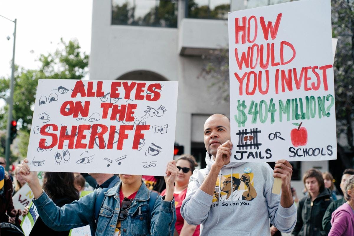 people at a rally holding signs that say "all eyes on the sheriff" and "how would you invest $404 million? jails or schools?"